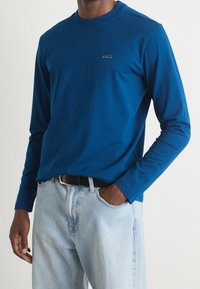 Långärmad blå t-shirt i bomull med rundad halsringning, med en liten "BOSS"-logotyp på bröstet. Matchas med ljusblå jeans.