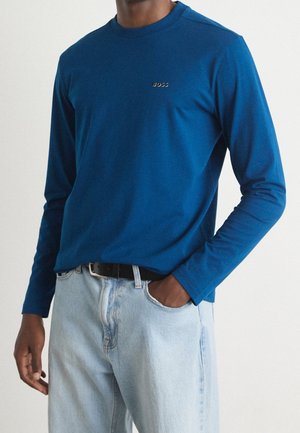Maglietta a maniche lunghe blu in cotone con scollo rotondo, caratterizzata da un piccolo logo "BOSS" sul petto. Abbinata a jeans azzurri.