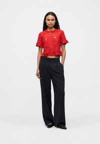 Červený krajkový crop top s volánkovými rukávy, kombinovaný s černými širokými kalhotami s vysokým pasem a černými špičatými balerínami. Jednoduché pozadí.