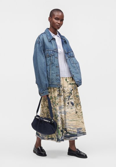 Personne portant une veste en jean bleue, une chemise blanche, une longue jupe imprimée, des mocassins noirs et tenant un sac à main bleu marine sur un fond uni.