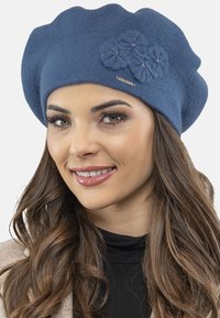 Beret tricotat albastru cu o suprafață texturată, având trei flori tricotate și un emblema metalică. Formă moale, rotundă, potrivită pentru diferite stiluri.