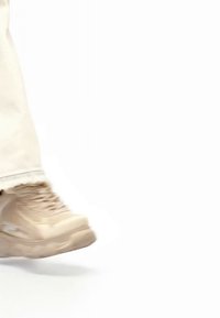 Pantof sport bej cu talpă groasă purtat cu pantaloni deschiși la culoare, prezentat în momentul mijlociu al mersului pe un fundal alb.