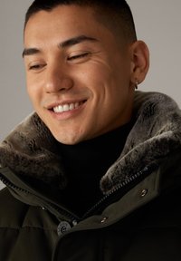 Glimlachende man met kort haar en een ringoorbel, gekleed in een donkergroene jas met een bontgekraagde kraag en een zwarte turtleneck tegen een effen achtergrond.