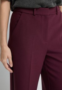 Burgundowe dopasowane spodnie o gładkiej fakturze, z bocznymi kieszeniami i płaskim przodem. Na nogawkach biegnie subtelne zagniecenie.