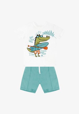 Weißes T-Shirt mit einem Cartoon-Krokodil in Sonnenbrille auf einem Paddleboard, kombiniert mit hellblauen Shorts. Bequemer Stoff, verspieltes Design.