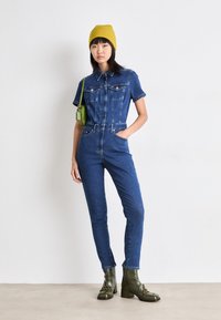 Denim overall in donkerblauw met korte mouwen, voorzakken en een getailleerde pasvorm. Gecombineerd met groene enkellaarzen en een gele beanie.