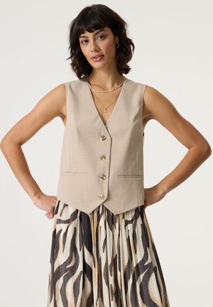 Femme aux cheveux courts et foncés portant un gilet beige boutonné et une jupe plissée aux rayures abstraites noires et marron, debout les mains sur les hanches.