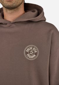 Brun hættetrøje med et cirkulært "Dolce Vita Pegador" logo i cremefarve. Stoffet ser blødt ud med en løs pasform og struktureret overflade.