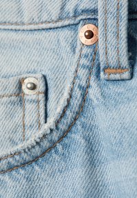 Gros plan sur un jean en denim bleu clair montrant la poche avant avec des rivets en cuivre et en argent ainsi que des détails de couture marron visibles.
