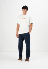 Man standing with one hand in pocket, wearing a white Tommy Jeans T-shirt, navy pants, and brown sneakers against a white background.