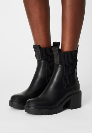 Botas cortas de cuero negro con un acabado de tela acanalada en la parte superior, tacón ancho y suela texturizada. Cuentan con una pestaña para facilitar su uso.