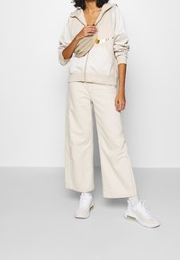 Beige zip-up huvtröja med fickor, kombinerad med vida krämfärgade byxor och vita sneakers med pastellfärgade detaljer. Midjeväska bärs över bröstet.