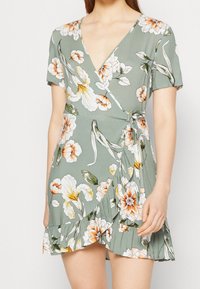 Kwiatowa sukienka w stonowanej zieleni, z dużymi białymi i pomarańczowymi kwiatami, krótkimi rękawami, głębokim dekoltem w kształcie litery V i falbanistym dołem.