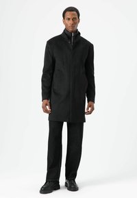 Zwarte wollen overjas met een hoge kraag, knoopsluiting aan de voorkant, zakken aan de zijkant en een gladde textuur, gecombineerd met donkere jeans en zwarte laarzen.