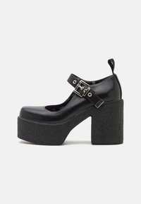 Black platform shoes with a rounded toe, featuring a wide textured heel and buckle strap. Smooth leather upper with a small back loop.