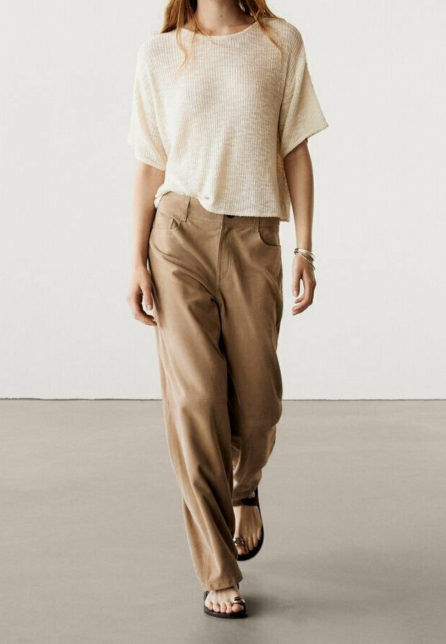 Beige tekstureret kortærmet top parret med løse, khaki-farvede bukser. Modellen bærer sorte sandaler, med en minimalistisk baggrund.