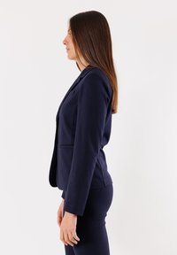 Blazer su misura di colore blu navy con chiusura a un bottone, tasche laterali e maniche lunghe, abbinato a pantaloni fitted coordinati. Tessuto morbido.