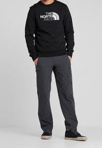 Černá mikina s kulatým výstřihem a bílým logem, kombinovaná se šedými cargo kalhotami, které mají kapsy a volný střih, nošena s tmavými botami.