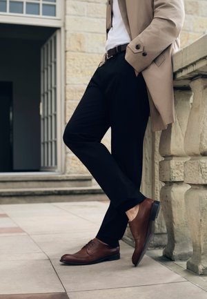 Brązowe skórzane buty typu oxford o gładkiej strukturze i zaokrąglonym nosku, w połączeniu z czarnymi spodniami i beżowym płaszczem, stojące na wyłożonej płytkami powierzchni.