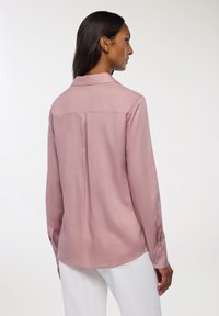 Camicia a maniche lunghe, simile alla seta, di un rosa tenue con una piega sul retro e colletto strutturato. Presenta polsini con bottoni e orlo curvato.
