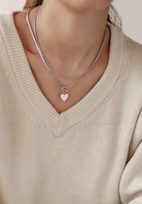 Zilveren ketting met gelaagde schakels en een hart bedel. Draagt over een lichtbeige gebreide trui met een V-hals ontwerp.