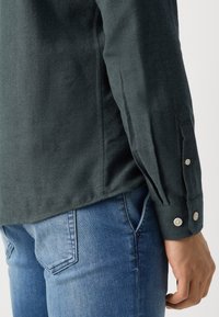 Dark green long-sleeve shirt with button cuffs, made of textured fabric. Paired with light blue jeans, showing side and arm details.