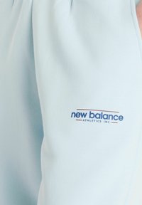 Pantalon de sport bleu clair avec une texture lisse, arborant un logo "New Balance" en bleu foncé et rouge sur le côté gauche.