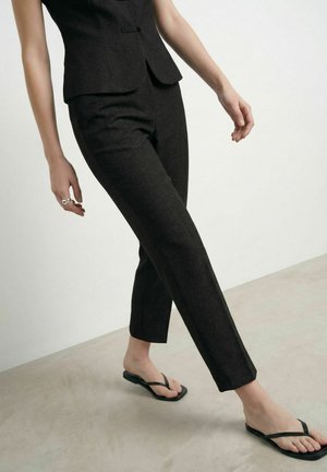 Personne portant un pantalon noir sur mesure et un blazer assorti, marchant pieds nus dans des tongs noires sur un sol clair.