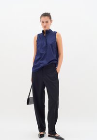 Bluză fără mâneci de culoare bleumarin, cu guler în picioare și placket cu butoane, asortată cu pantaloni negri ajustați și pantofi negri fără toc. Geantă compactă de mână de culoare neagră.