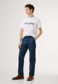 Camiseta blanca con el logotipo "PEPE JEANS" en negro y rojo, combinada con pantalones azul oscuro. El modelo lleva zapatos de ante marrones, de pie sobre un fondo claro.