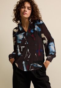 Lange mouwen blouse in donkere stof met abstracte blauwe, rode en crèmekleurige patronen; heeft een V-hals en licht geplooide mouwen.