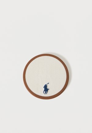 Στρογγυλή μπεζ υφασμάτινη επιφάνεια με καφέ δερμάτινη περίμετρο, που διαθέτει μικρό σήμα με κέντημα μπλε σκούρου παίκτη πόλο στο κάτω κέντρο.