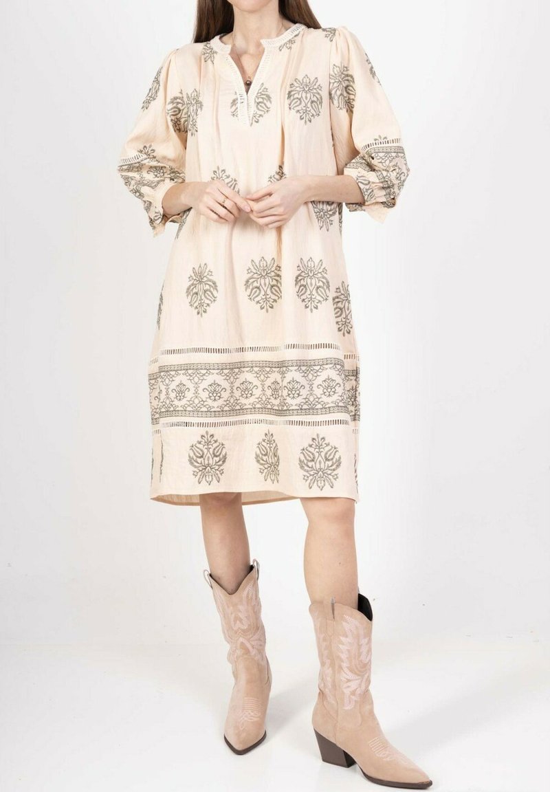 Robe rose pâle avec une broderie florale grise complexe, des manches froncées et un col en V, associée à des bottes de cowboy beiges.