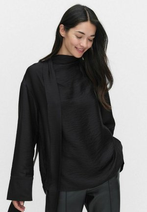 Sort, tekstureret langærmet top med høj hals og brede manchetter, parret med sorte bukser. Stoffet ser blødt og let krøllet ud.