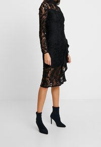 Robe en dentelle noire avec un col haut, des manches longues et un détail de volant à l'ourlet. Portée avec des bottines noires à bout pointu.