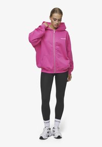 Roza zip-up dukserica s prednjim džepom, kombinirana s crnim tajicama. Model nosi bijele čarape i tenisice, stilizirano s kapuljačom na sredini.