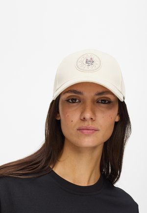 Γυναίκα με μακριά σκούρα μαλλιά που φοράει ένα λευκό καπέλο μπέιζμπολ με έμβλημα γάτας και μαύρο πουκάμισο, κοιτάζοντας ευθεία μπροστά με ουδέτερη έκφραση.