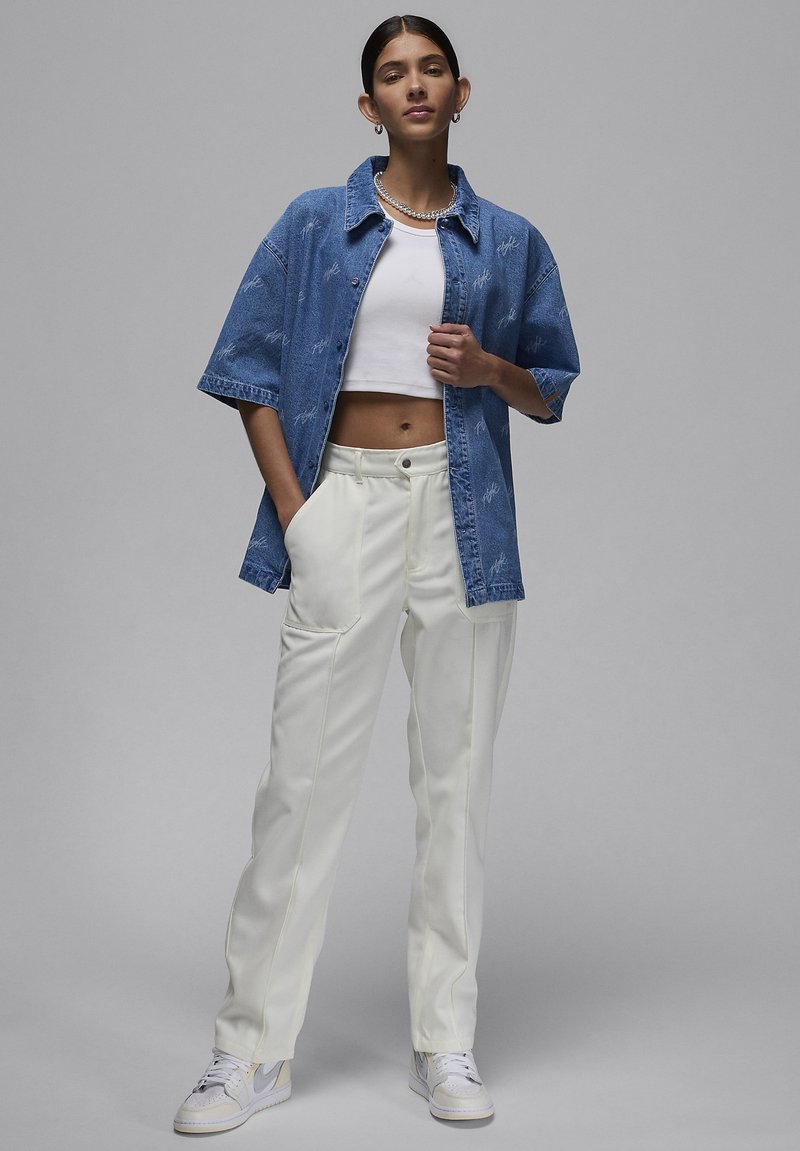Kobieta ubrana w niebieską, oversize'ową dżinsową kurtkę, białą bluzkę crop top, białe spodnie z wysokim stanem i białe trampki, stojąca na szarym tle.