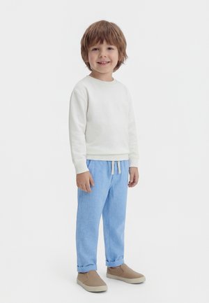 Junger Junge lächelt, trägt ein weißes Langarmshirt, hellblaue Hosen mit Kordelzug und beige Schlupfschuhe, steht vor einem weißen Hintergrund.