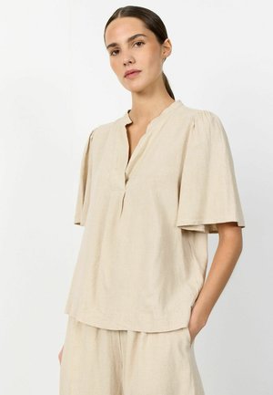 Frau mit dunklen Haaren zu einem niedrigen Pferdeschwanz gebunden, trägt eine beige kurzärmelige V-Ausschnitt-Bluse und passende Hose, steht vor weißem Hintergrund.