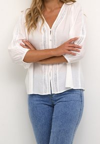 Blusa blanca de botones con encaje en los bordes y mangas de tres cuartos, combinada con jeans ajustados azules. Tejido texturizado, ajuste relajado.