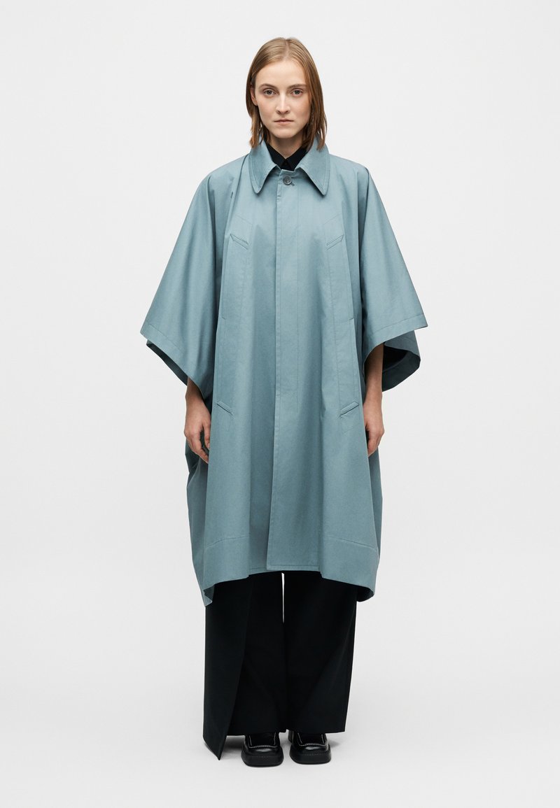 Manteau ample bleu clair avec col boutonné, manches courtes, détails minimalistes et silhouette droite, associé à un pantalon noir.