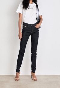 Mörka denim skinny jeans i kombination med en vit bomullströja med en logotyp, kompletterad med en svart axelväska och beige klackar.