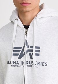 Lichtgrijze zip-up hoodie met trekkoorden, voorzien van een donkergrijs logo en de tekst "ALPHA INDUSTRIES" gedrukt op de voorkant. Zachte stoftextuur.