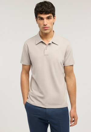 Junger Mann mit kurzen lockigen Haaren, der ein beiges Poloshirt mit kurzen Ärmeln und marineblaue Hosen trägt, steht mit einer Hand in der Tasche.