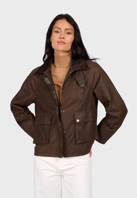 Hnědá kožená bunda se zapínáním na zip, dvěma velkými předními kapsami a kostkovanou podšívkou. Obsahuje kontrastní hnědý límec a volný střih.