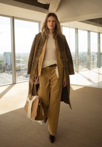 Woman walking indoors wearing layered brown suede and corduroy coat and pants, carrying beige handbag, with cityscape visible through floor-to-ceiling windows.