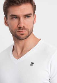 Maglietta bianca a V in cotone. Presenta un piccolo logo nero sul lato sinistro del petto. Tessuto morbido e design aderente.