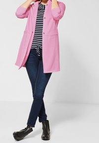 Vaaleanpunainen pitkä neuletakki, jossa on kaksinkertaiset napit, yhdistettynä raidalliseen paitaan ja tummiin slim fit -farkkuihin. Mustat nilkkurit viimeistelevät asun.