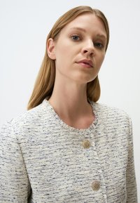 Giacca in tweed bianca e blu con texture, dal collo rotondo, caratterizzata da due bottoni dorati e bordi sfrangiati.